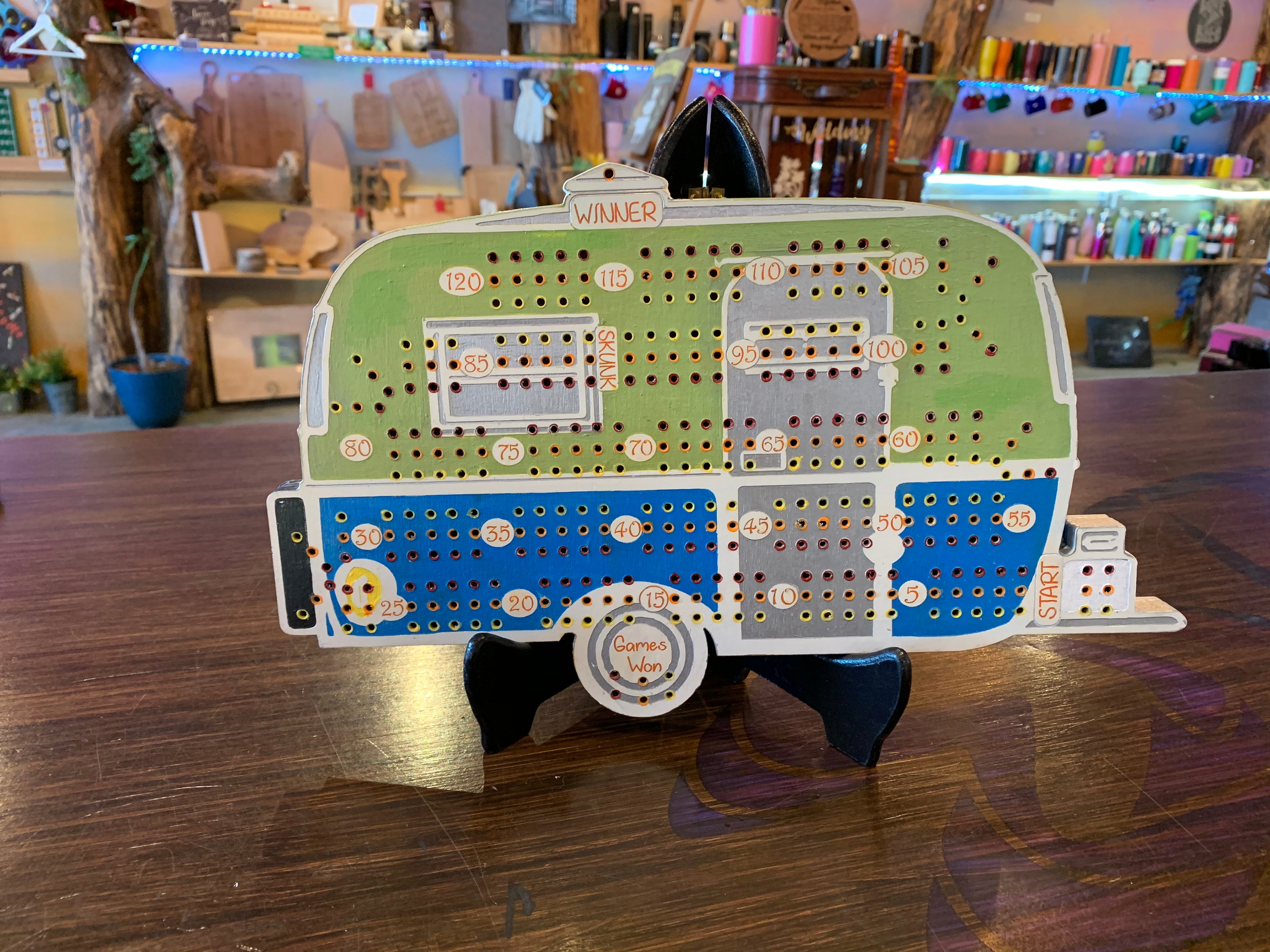Cribbage board shaped like a camper on a wooden floor with a store in the background
